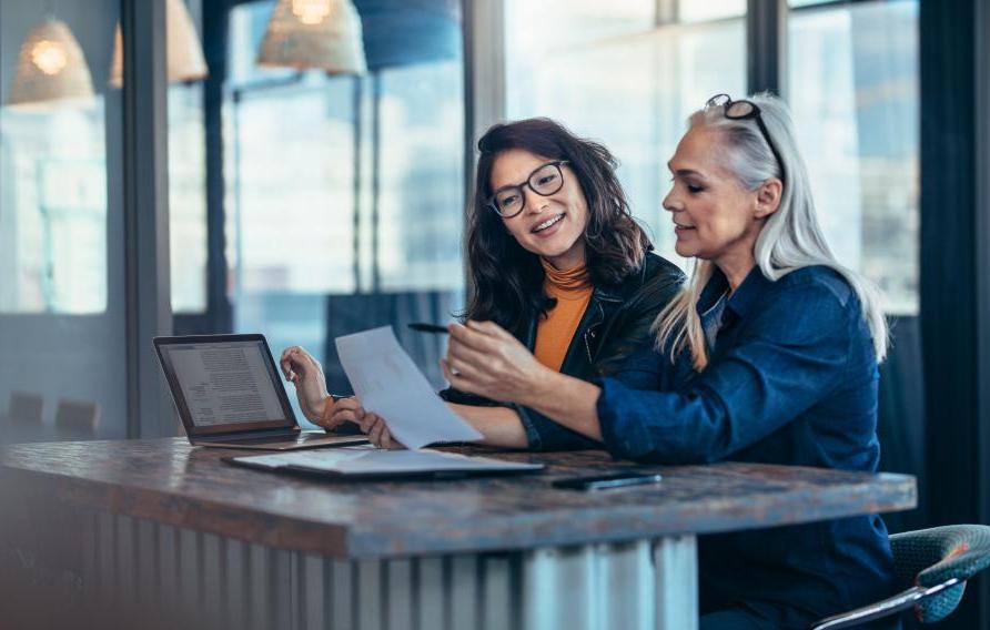 Two women analyzing documents while sitting on a table in office. Woman executives at work in office discussing some paperwork.