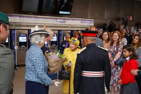 Her Majesty the Queen dressed in yellow coat and hat, with a school girls in red sweater, a man in blue/red uniform, a lady in blue with hat and other members of the public.